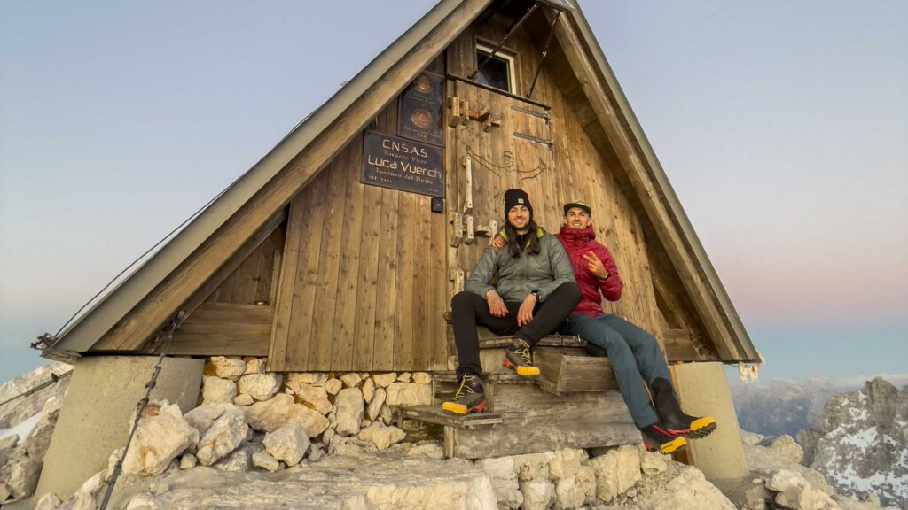 Due escursionisti seduti davanti a un bivacco in montagna al tramonto.